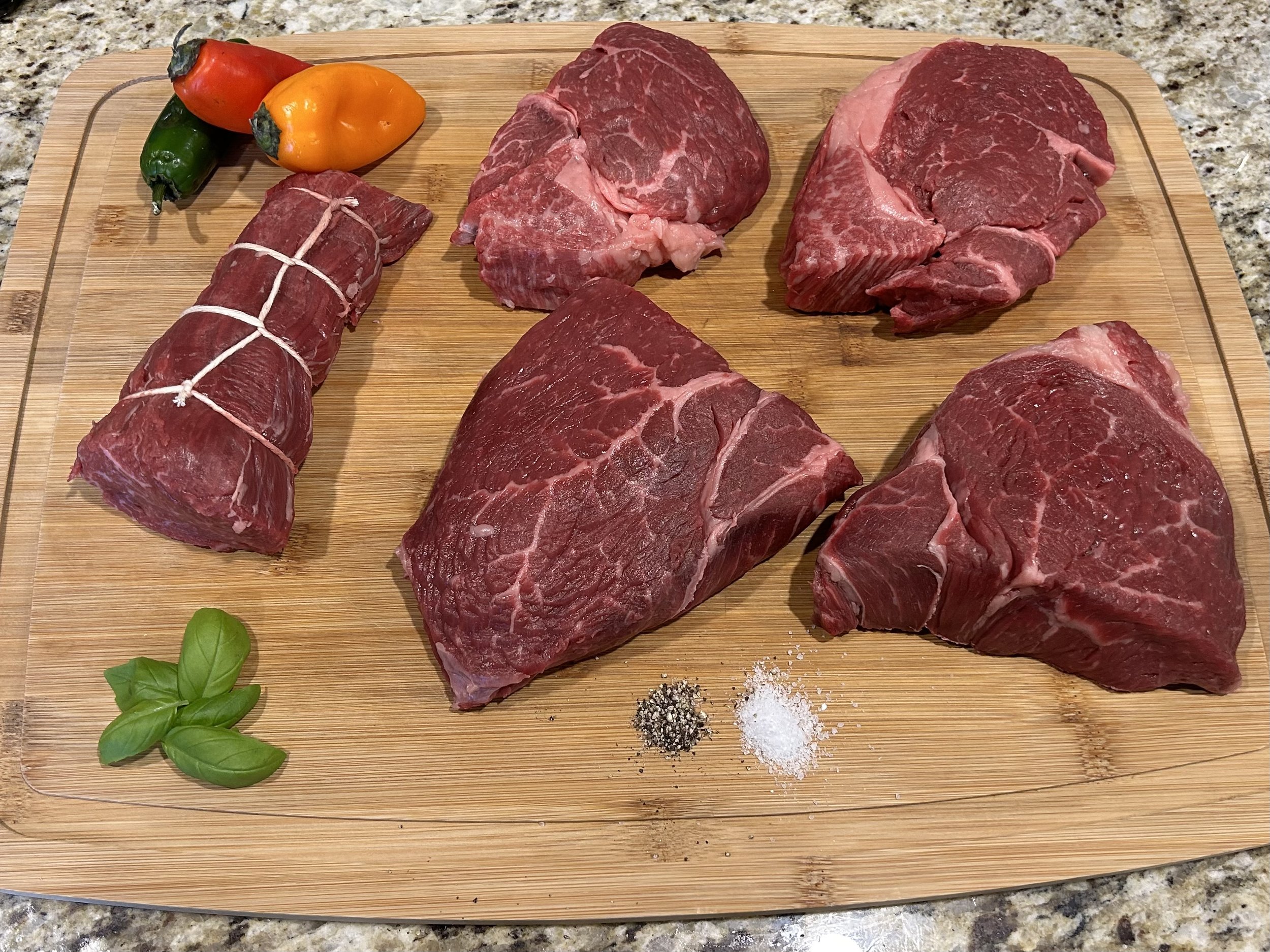 Jake+Moore+-+Prodcuts_Page_ShopAll Assorted cuts of raw beef on a wooden cutting board, including a tied roast, accompanied by fresh basil leaves and colorful peppers, with salt and pepper for seasoning.