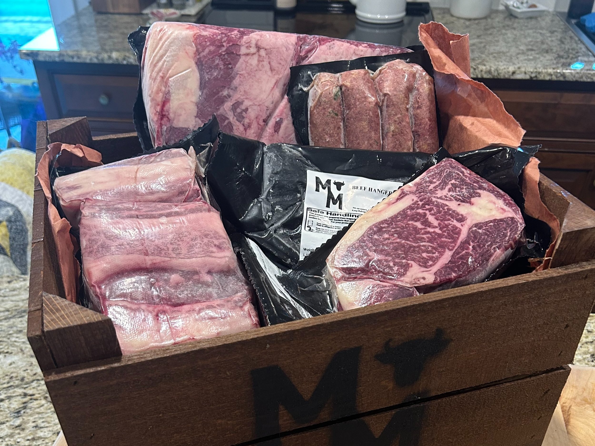 Jake+Moore+-+Products_Page_Bundles A wooden crate filled with various cuts of beef, including marbled steaks, sausages, and beef short ribs, all packaged in plastic and butcher paper. The crate features a cow logo, highlighting premium quality meat for grilling and culinary use.