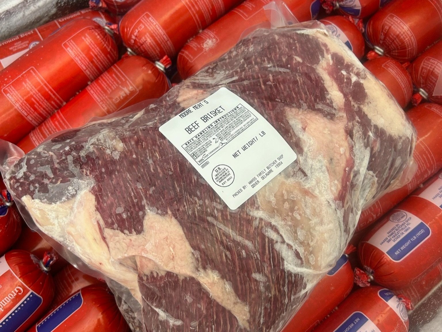 Jake+Moore+-+Products_Page_Half_Steer Vacuum-sealed beef brisket packaged with a label indicating net weight, surrounded by red packages of ground beef sausages. Ideal for barbecues and slow cooking.