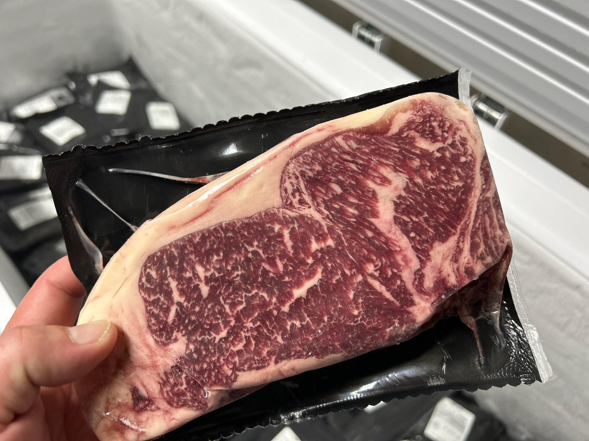 Jake+Moore+-+Products_Quater_Steer Close-up of a hand holding a vacuum-sealed package containing a marbled steak, showcasing its rich texture and quality. In the background, a freezer filled with additional black-packaged meat products is visible. Ideal for gourmet cooking and grilling enthusiasts.
